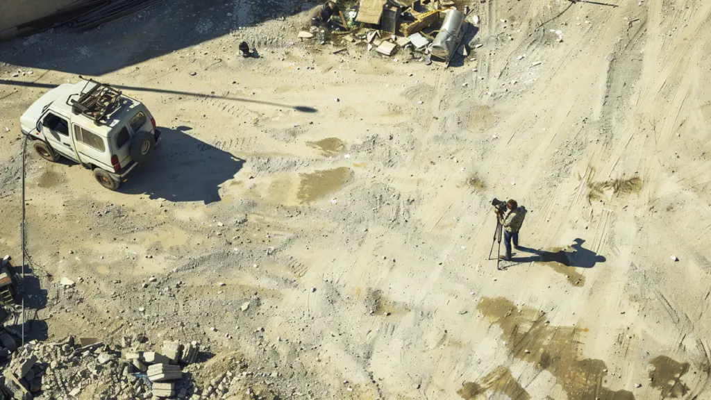 Overhead view of a person with camera equipment on a dusty site beside a white vehicle.